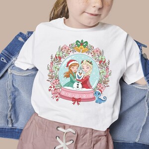 May include: A white t-shirt featuring a graphic of two characters with braided hair and a snowman inside a snow globe. The globe is surrounded by a floral wreath with golden bells at the top and a small blue lizard wearing a Santa hat nearby.