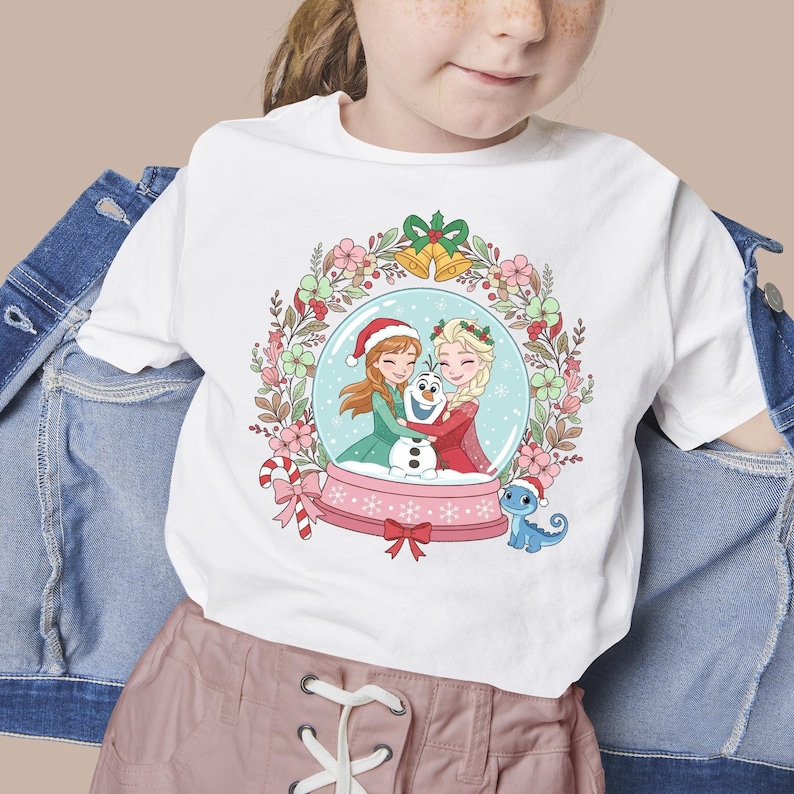 May include: A white t-shirt featuring a graphic of two characters with braided hair and a snowman inside a snow globe. The globe is surrounded by a floral wreath with golden bells at the top and a small blue lizard wearing a Santa hat nearby.
