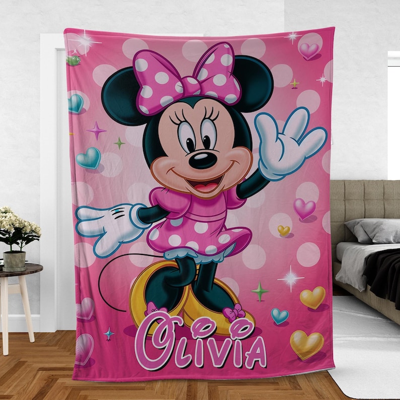 Puede incluir: Una manta de forro polar rosa con Minnie Mouse con un vestido y lazo de lunares rosas. La manta tiene un fondo rosa con lunares blancos, corazones y el nombre "Olivia" en cursiva.