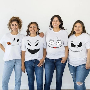 May include: Four white short-sleeve t-shirts featuring black graphic prints of character faces. The first shirt shows a ghost with a small orange pumpkin, the second a jagged grin, the third a stitched face with red lips, and the fourth a wide smile.