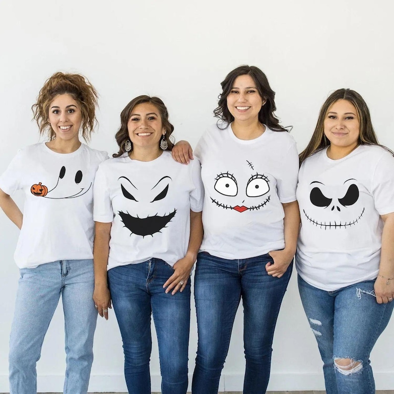 May include: Four white short-sleeve t-shirts featuring black graphic prints of character faces. The first shirt shows a ghost with a small orange pumpkin, the second a jagged grin, the third a stitched face with red lips, and the fourth a wide smile.