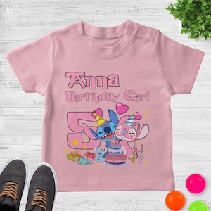 May include: A light pink short-sleeved t-shirt featuring a graphic of two blue and pink cartoon characters wearing party hats. The text reads Anna Birthday Girl above a large number 5, a birthday cake, gifts, and small yellow stars.