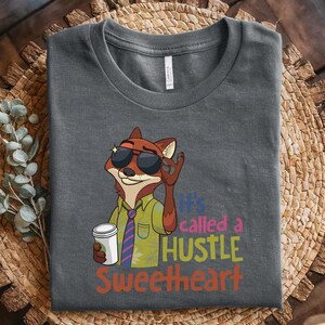 May include: A dark gray t-shirt featuring a cartoon fox wearing sunglasses, a green shirt, and a striped tie, holding a white coffee cup. The text reads, "it's called a HUSTLE SWEETHEART" in colorful, stacked lettering next to the fox illustration.