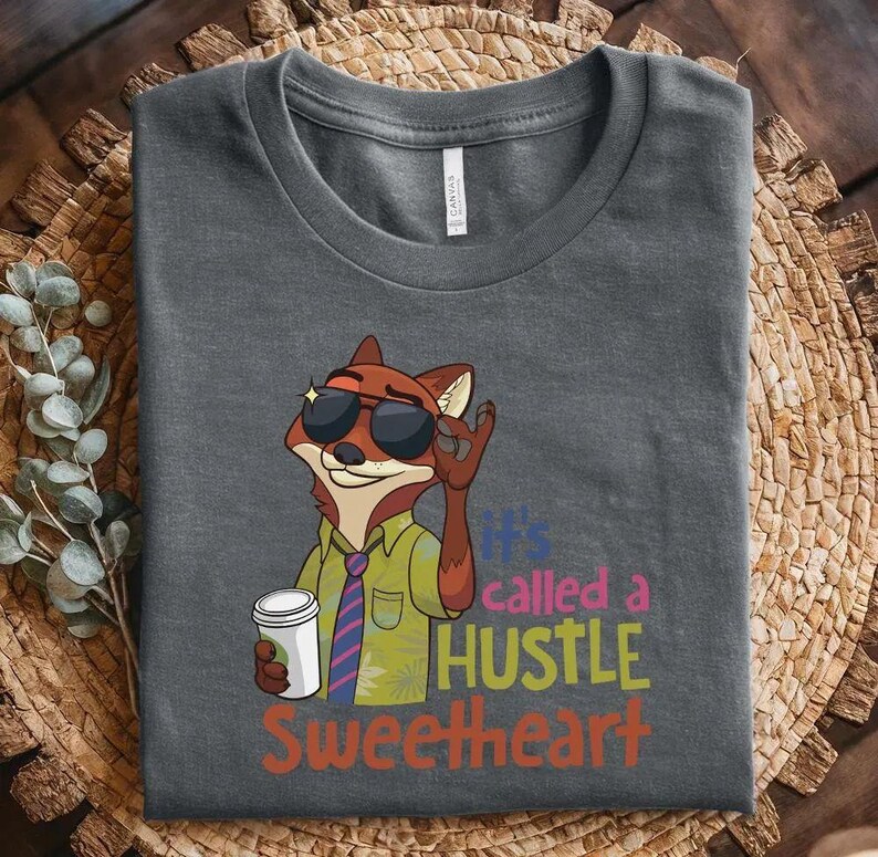 May include: A dark gray t-shirt featuring a cartoon fox wearing sunglasses, a green shirt, and a striped tie, holding a white coffee cup. The text reads, "it's called a HUSTLE SWEETHEART" in colorful, stacked lettering next to the fox illustration.