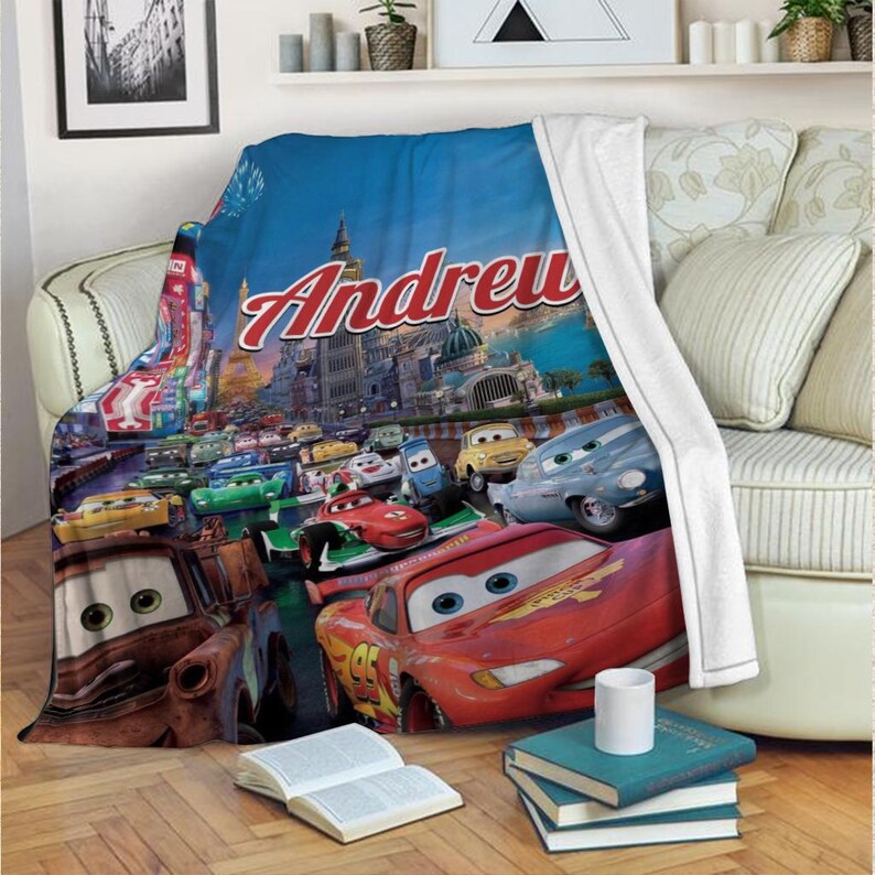 Op de afbeelding: Een zachte deken met een kleurrijke sc&egrave;ne uit de animatiefilm Cars, met de naam "Andrew" in rode letters. De deken toont Bliksem McQueen en andere personages die racen tegen een stadsgezicht.