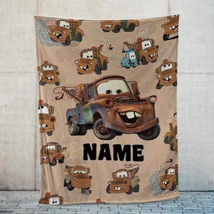 May include: A tan blanket featuring a repeating pattern of a cartoon tow truck character. The central image is a large version of the character with the word "NAME" in black block letters below it. The blanket is made of a soft material.