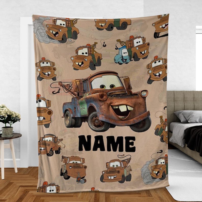 May include: Beige blanket featuring a repeating pattern of cartoon tow trucks in various poses. A large, rusty brown tow truck is centered, with the word "NAME" in black below. The blanket is likely made of soft fabric.