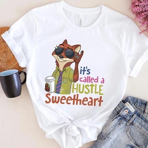 May include: A white short-sleeved t-shirt featuring a graphic of a cartoon fox wearing sunglasses and a green shirt with a tie, holding a white cup. The text reads, "it's called a HUSTLE SWEETHEART" in colorful, stylized lettering.