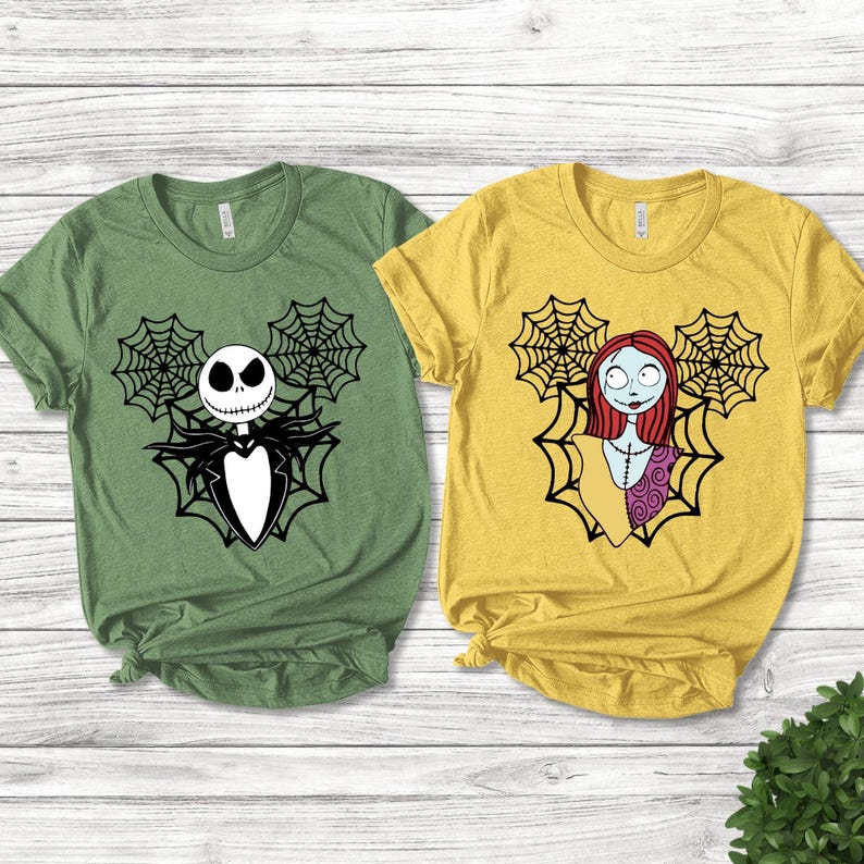 K&ouml;nnte beinhalten: Ein salbeigr&uuml;nes T-Shirt mit einer schwarzen Grafik eines Skelett-Charakters im Anzug mit Spinnenweben und ein senfgelbes T-Shirt mit einer schwarzen Grafik eines weiblichen Charakters mit roten Haaren und Patchwork-Kleid mit Spinnenweben.