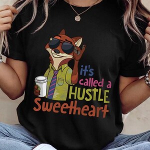 May include: A black t-shirt featuring a cartoon fox wearing sunglasses, a green short-sleeved shirt, and a striped tie. The fox holds a white coffee cup. The text reads: it's called a HUSTLE SWEETHEART in blue, pink, yellow, and orange lettering.