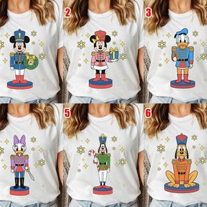 May include: A white t-shirt featuring a graphic of a Disney character dressed as a nutcracker standing on a circular base, surrounded by yellow snowflake icons. The characters include Mickey, Minnie, Donald, Daisy, Goofy, and Pluto in festive attire.