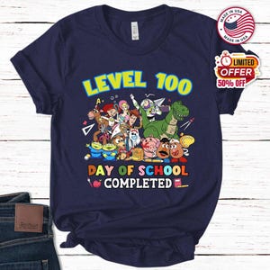 May include: A navy blue short-sleeve t-shirt featuring a colorful graphic of various animated toy characters. The text reads LEVEL 100 DAY OF SCHOOL COMPLETED, accompanied by illustrations of school supplies like a ruler, scissors, and an apple.