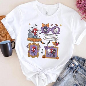 May include: A white short-sleeve t-shirt featuring a colorful Halloween-themed graphic print of cartoon characters including Spider-Man, Miles Morales, and Ghost-Spider. The design includes pumpkins, spiderwebs, bats, and text reading Spidey and Amazing Spider-Man.