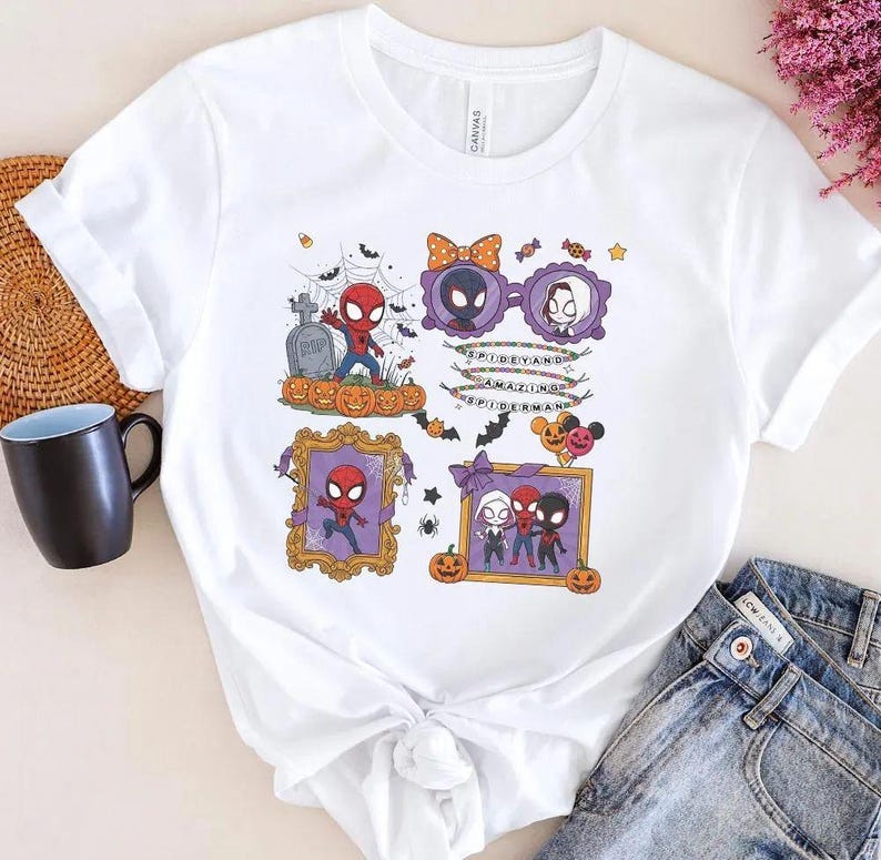 May include: A white short-sleeve t-shirt featuring a colorful Halloween-themed graphic print of cartoon characters including Spider-Man, Miles Morales, and Ghost-Spider. The design includes pumpkins, spiderwebs, bats, and text reading Spidey and Amazing Spider-Man.