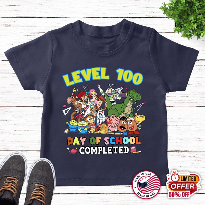 May include: A navy blue short-sleeve t-shirt featuring a colorful graphic of various animated toy characters. The text reads LEVEL 100 DAY OF SCHOOL COMPLETED, accompanied by illustrations of school supplies like a pencil, an apple, and a ruler.