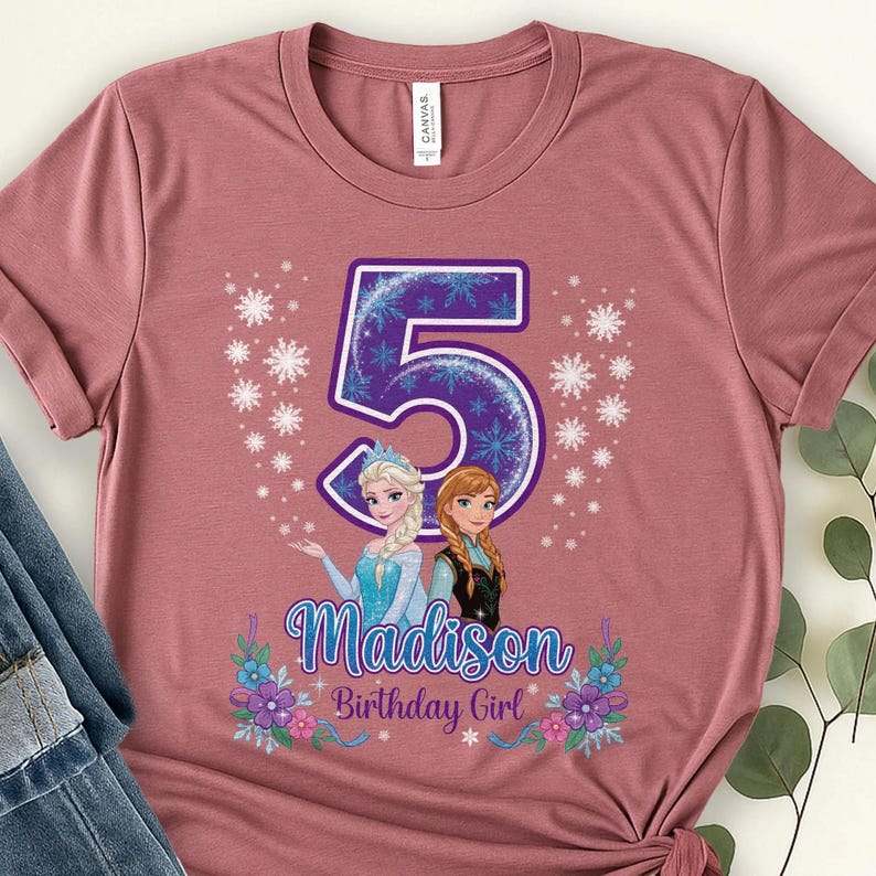 以下が含まれることがあります： くすんだピンク色のクルーネックTシャツ。雪の結晶模様が描かれた大きな紫色の数字の5が中央にあります。その下には、金髪と赤褐色の髪のキャラクターのイラストが描かれています。テキストにはMadison Birthday Girlと記されています。