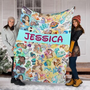 K&ouml;nnte beinhalten: Eine farbenfrohe Decke mit Zeichentrickfiguren und dem Namen "JESSICA" in rosa Buchstaben auf einem hellblauen Banner. Die Decke wird von zwei Personen gehalten. Die Decke hat einen wei&szlig;en Hintergrund mit verschiedenen Zeichentrickfiguren.