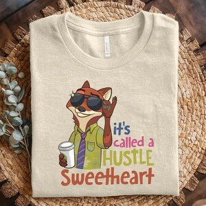 May include: A beige crew neck t-shirt featuring a cartoon fox wearing sunglasses, a green shirt, and a striped tie, holding a white coffee cup. The text reads, it's called a HUSTLE SWEETHEART, in blue, pink, green, and orange lettering.
