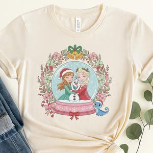May include: A cream-colored short-sleeve t-shirt featuring a graphic of two animated characters hugging a snowman inside a snow globe. The globe is surrounded by a floral wreath with bells, and a small blue lizard wearing a Santa hat sits nearby.