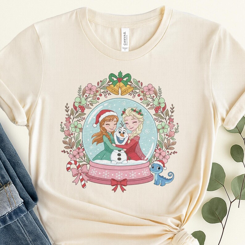May include: A cream-colored short-sleeve t-shirt featuring a graphic of two animated characters hugging a snowman inside a snow globe. The globe is surrounded by a floral wreath with bells, and a small blue lizard wearing a Santa hat sits nearby.