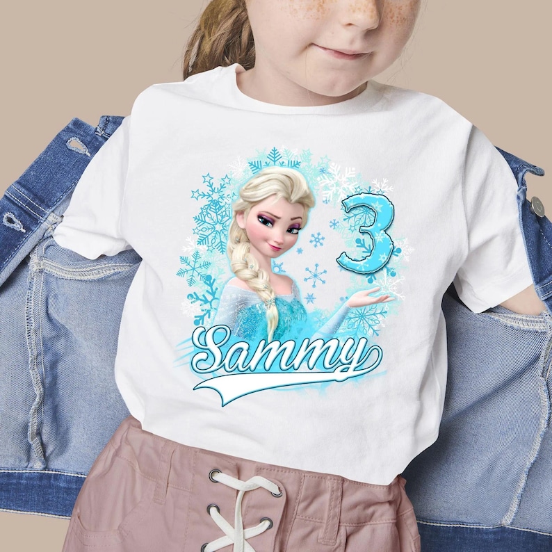 May include: A white t-shirt featuring a graphic of a blonde character with a braid in a blue dress, surrounded by light blue snowflakes. The design includes the large blue number 3 and the name Sammy in a cursive script font below the character.