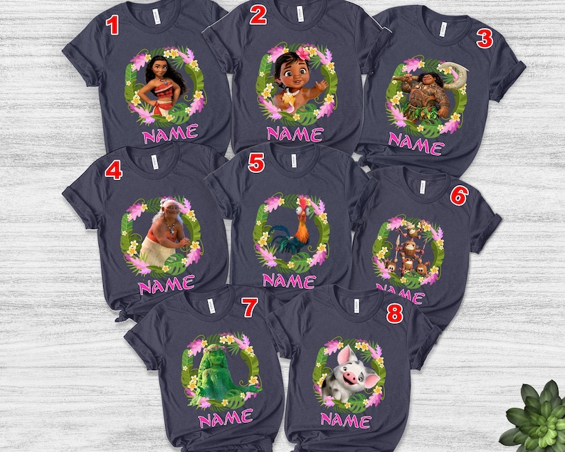 May include: Eight dark gray short-sleeve t-shirts featuring various Moana character graphics inside floral and leaf wreaths. Each shirt includes the word NAME in pink text below the graphic and a numbered red tag in the top corner.