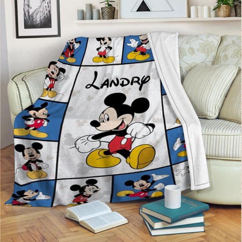 Puede incluir: Una manta blanca y azul con Mickey Mouse en varias poses. La manta tiene un dise&ntilde;o de cuadr&iacute;cula con im&aacute;genes de Mickey Mouse sobre un fondo azul. El nombre "Landry" est&aacute; escrito en cursiva en la manta.