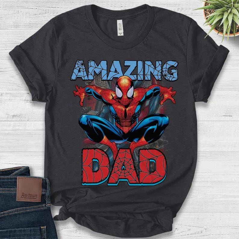 Pu&ograve; includere: Una t-shirt nera a maniche corte con una grafica del personaggio Spider-Man in posizione accovacciata. Sopra il personaggio c'&egrave; la parola AMAZING in azzurro, e sotto la parola DAD in rosso con un motivo a ragnatela sovrapposto.