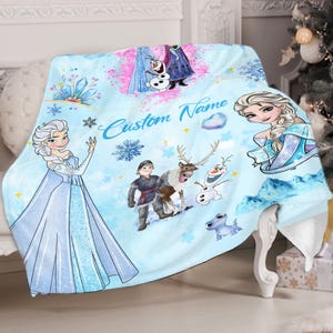 May include: A blue and white fleece blanket with a Frozen theme. It features characters like Elsa, Anna, Olaf, and Kristoff. The blanket has the text "Custom Name" printed on it.