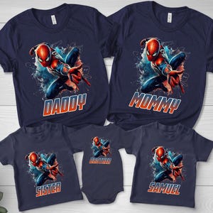 May include: A collection of five navy blue t-shirts and one bodysuit featuring a graphic of a red and blue superhero in a dynamic pose. Each garment displays the character above text that reads DADDY, MOMMY, SISTER, BROTHER, or SAMUEL.