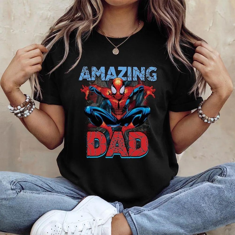Pu&ograve; includere: Una t-shirt nera a maniche corte con una grafica del supereroe Spider-Man in posizione accovacciata. Sopra il personaggio si trova la parola AMAZING in azzurro, e sotto la parola DAD in rosso con un motivo a ragnatela nero.
