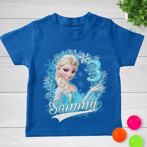 May include: A bright blue short-sleeved t-shirt featuring a graphic of a blonde character with a braid, surrounded by a snowflake wreath. The design includes the number 3 in a light blue font and the name Sammy written in white script below.