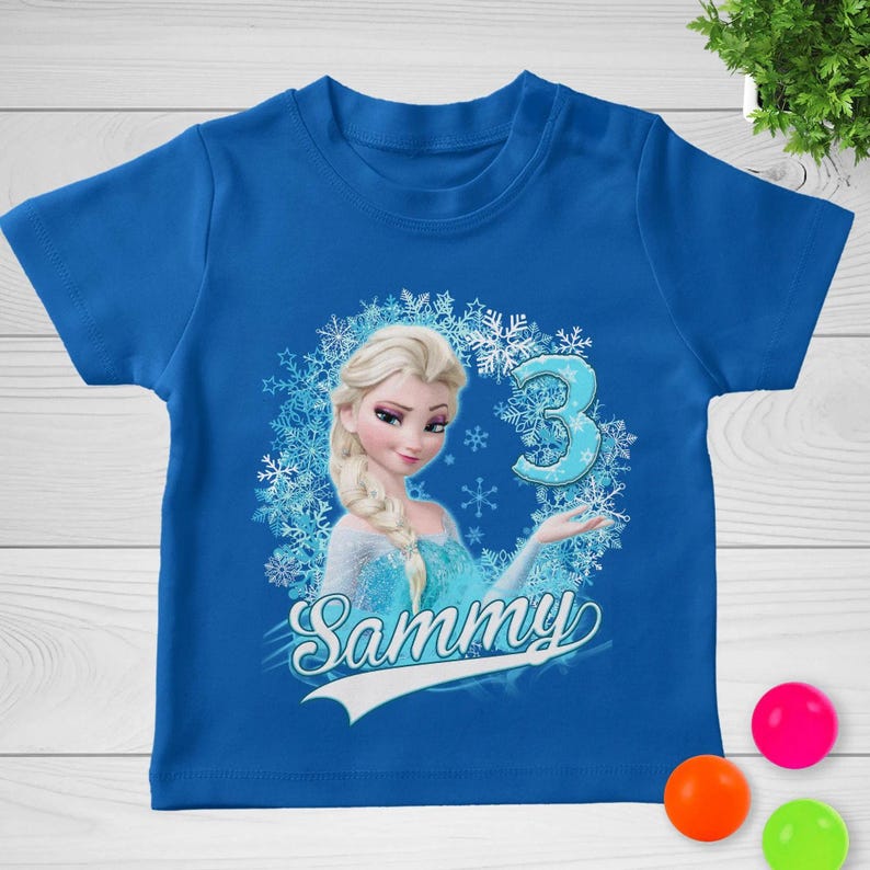 May include: A bright blue short-sleeved t-shirt featuring a graphic of a blonde character with a braid, surrounded by a snowflake wreath. The design includes the number 3 in a light blue font and the name Sammy written in white script below.