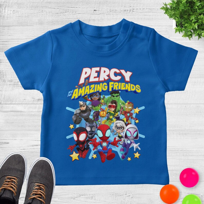 May include: A bright blue short-sleeve t-shirt featuring a graphic print of various cartoon superheroes. The text PERCY and his AMAZING FRIENDS is printed in large, bold letters above the characters, which include figures in red, green, and white.