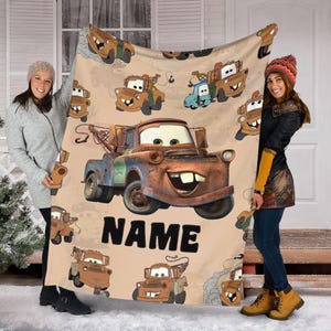 May include: A beige blanket featuring a cartoon illustration of the character Mater from the movie Cars. The blanket has a repeating pattern of Mater in various poses, with the word "NAME" printed in large, bold letters.