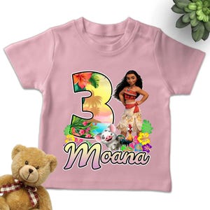 May include: A light pink short-sleeved t-shirt featuring a large number three with a tropical sunset and floral pattern. The graphic includes the character Moana, Pua the pig, and Heihei the rooster, with the name Moana printed in cursive below.