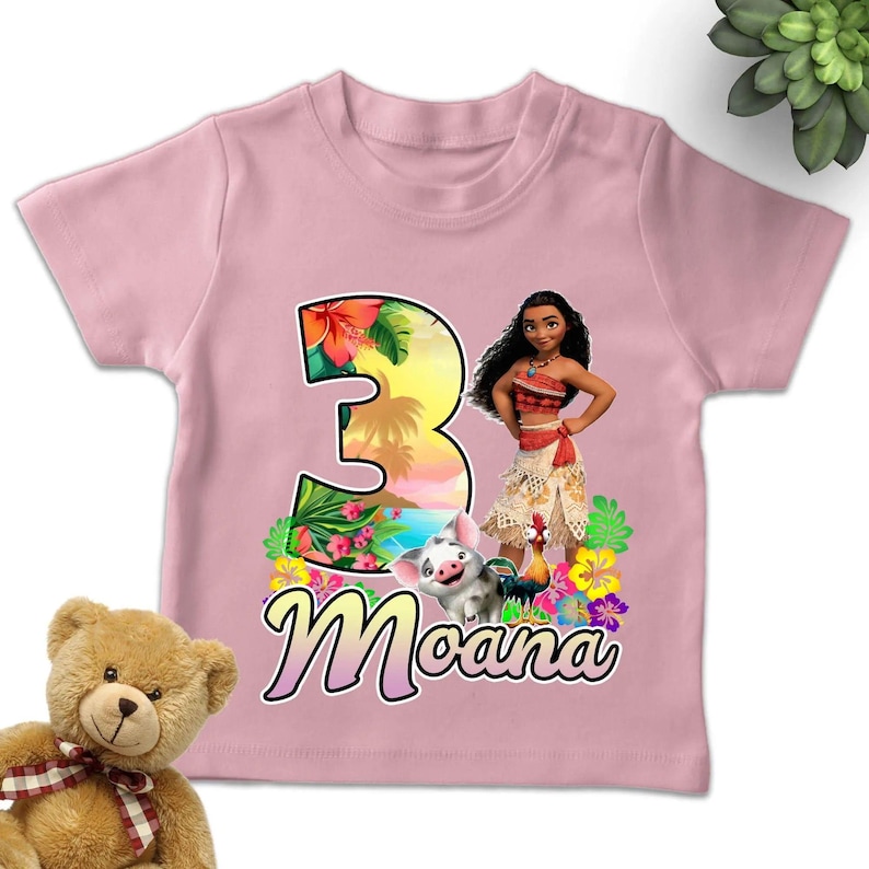 May include: A light pink short-sleeved t-shirt featuring a large number three with a tropical sunset and floral pattern. The graphic includes the character Moana, Pua the pig, and Heihei the rooster, with the name Moana printed in cursive below.