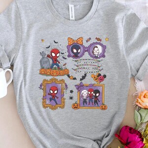 May include: A heather gray t-shirt featuring a Halloween-themed graphic with Spider-Man characters, pumpkins, a tombstone, and spiderwebs. The design includes text that reads SPIDEY AND AMAZING SPIDERMAN, surrounded by bats and decorative frames.