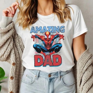 Pu&ograve; includere: Una t-shirt bianca a maniche corte con la grafica di un supereroe rosso e blu in posizione accovacciata. Sopra la figura si trova la parola AMAZING in blu, e sotto la parola DAD in grandi lettere rosse con un motivo a ragnatela nero.