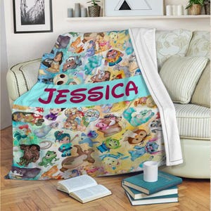 K&ouml;nnte beinhalten: Eine farbenfrohe Decke mit verschiedenen Zeichentrickfiguren. Die Decke hat ein hellblaues Band mit dem Namen "JESSICA" in gro&szlig;en, rosa Buchstaben. Die Decke ist &uuml;ber ein Sofa drapiert.