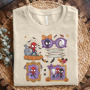 May include: A beige t-shirt featuring a graphic print of various Spider-Man characters in a Halloween theme. The design includes pumpkins, a tombstone, spiderwebs, bats, and frames with text that reads SPIDEY AND AMAZING SPIDERMAN.