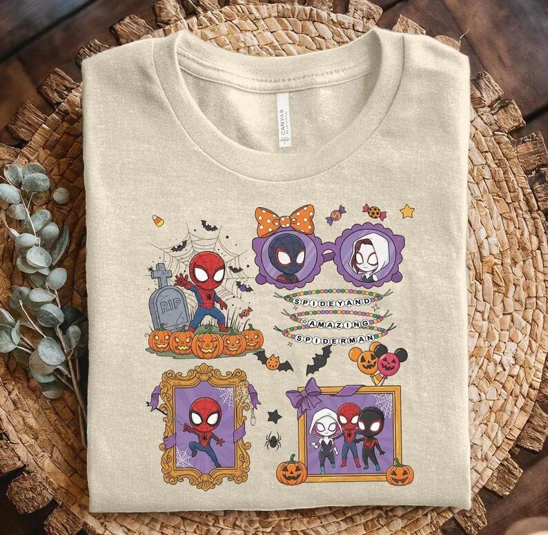 May include: A beige t-shirt featuring a graphic print of various Spider-Man characters in a Halloween theme. The design includes pumpkins, a tombstone, spiderwebs, bats, and frames with text that reads SPIDEY AND AMAZING SPIDERMAN.