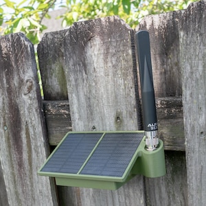 May include: An olive green solar-powered device featuring a rectangular solar panel and a vertical black antenna with the text "ALFA 915" printed on the base. The unit is mounted to a weathered wooden fence, providing a compact and functional design.