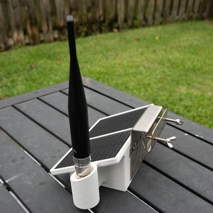 May include: A white 3D-printed mount featuring a black vertical antenna, a small rectangular solar panel, and a metallic bracket secured with two wing nuts. The device is designed for external mounting and includes a cylindrical base for the antenna.