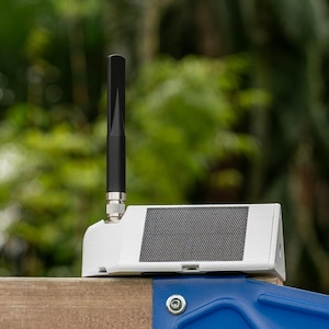 May include: A white, wedge-shaped electronic device featuring a black vertical antenna and a rectangular solar panel on its slanted top surface. The unit is mounted on a wooden structure with a blue metal bracket visible at the base.