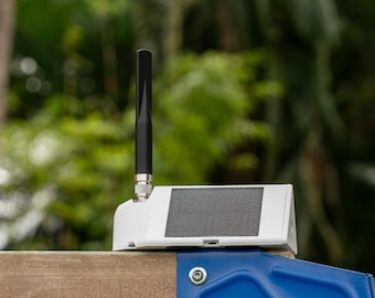 May include: A white, wedge-shaped electronic device featuring a black vertical antenna and a rectangular solar panel on its slanted top surface. The unit is mounted on a wooden structure with a blue metal bracket visible at the base.