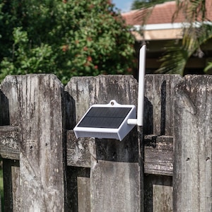 May include: A white solar panel device with a rectangular black solar cell array and a vertical white antenna attached to the side. The unit features a small mounting hole at the top center and is constructed from a solid white plastic housing.