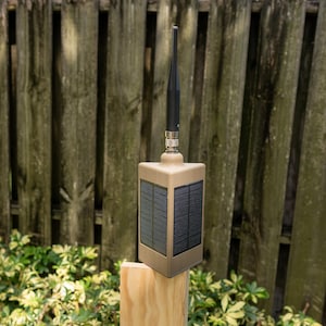 May include: A tan, rectangular electronic device featuring solar panels on its sides and a black antenna extending from the top. The unit is mounted on a light-colored wooden post, showcasing a functional design for outdoor signal transmission.