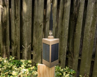 Puede incluir: Un dispositivo electrónico rectangular de color tostado con paneles solares en los laterales y una antena negra que sobresale por la parte superior. La unidad está montada sobre un poste de madera clara, mostrando un diseño funcional.