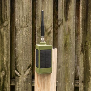 May include: A rectangular olive green electronic device featuring a black solar panel on its front face and a vertical black antenna extending from the top center. The unit is mounted on a light-colored wooden post with a smooth, finished surface.